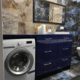 Niebieska szafka łazienkowa z szufladami i blatem, z wbudowaną pralką, lustro w czarnej ramie i marmurowe płytki na ścianach i podłodze.