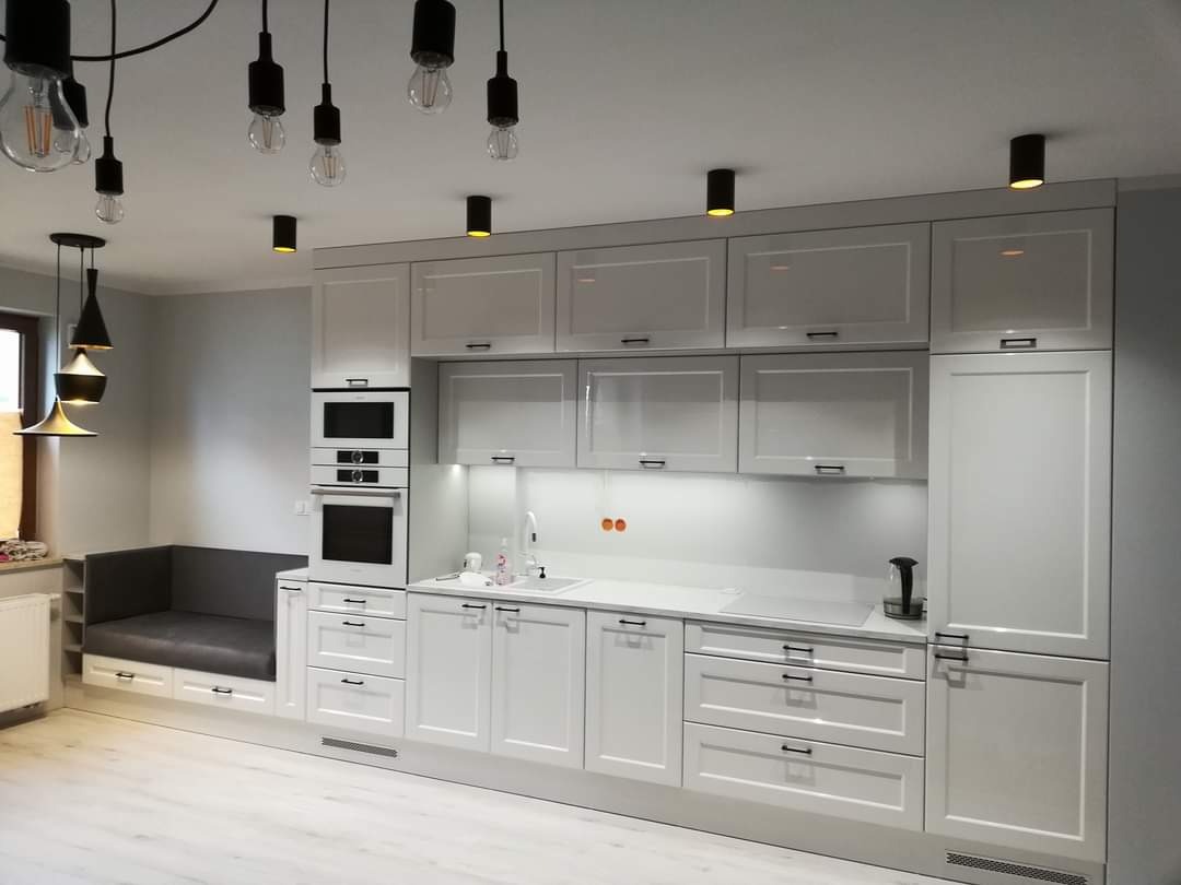 Jasna kuchnia z białymi frontami, czarnymi uchwytami i oświetleniem punktowym, z zabudowaną strefą wypoczynkową z szarą tapicerką.