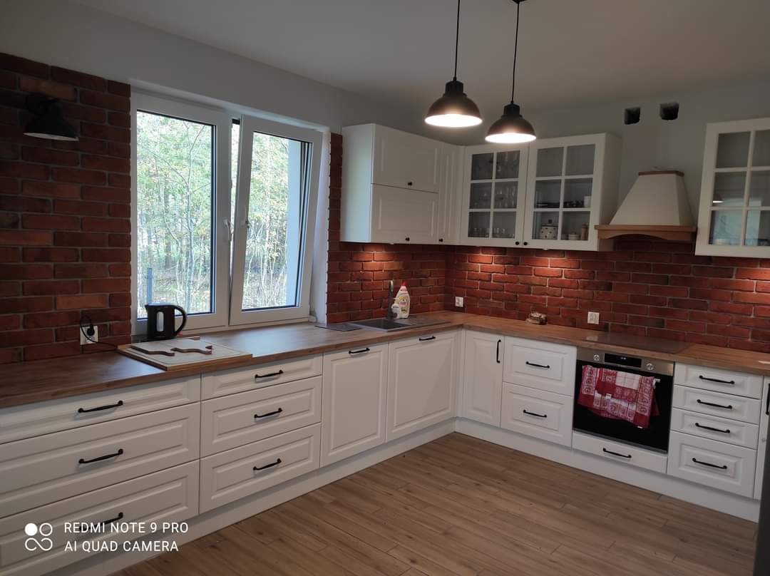 Biała kuchnia w stylu rustykalnym z drewnianymi blatami, ceglaną ścianą i czarnymi uchwytami, oświetlona wiszącymi lampami.