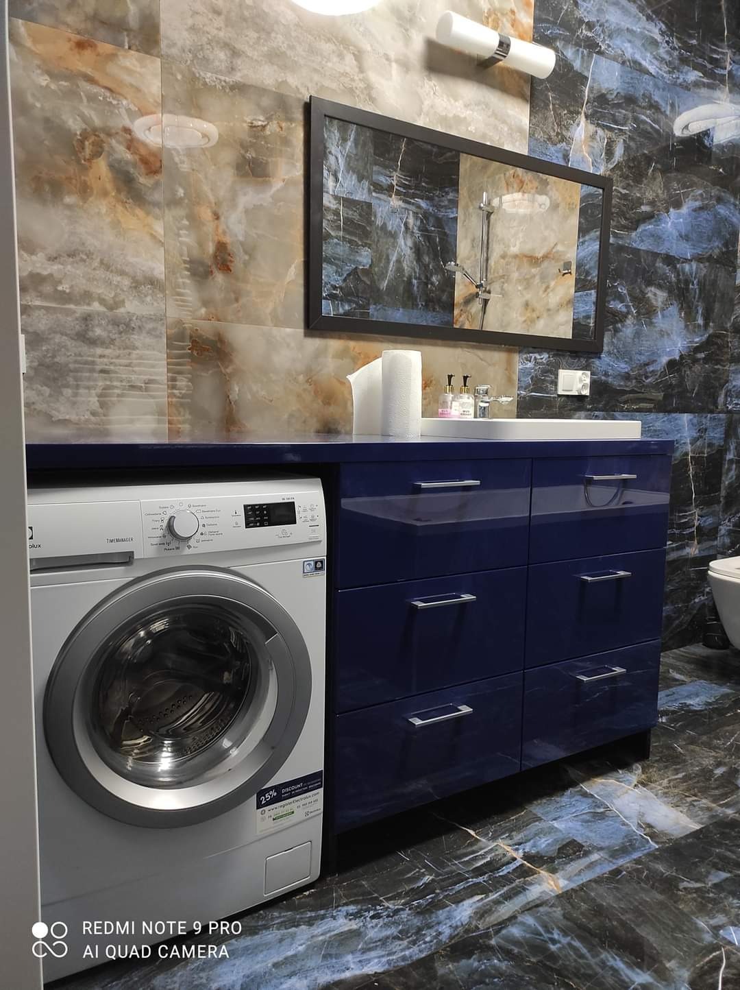 Niebieska szafka łazienkowa z szufladami i blatem, z wbudowaną pralką, lustro w czarnej ramie i marmurowe płytki na ścianach i podłodze.