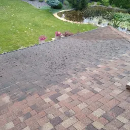 Częściowo wyczyszczony dach pokryty gontem bitumicznym, z widocznym ogrodem, trawnikiem i stawem w tle.