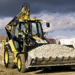 Żółto-czarna koparko-ładowarka CAT z pełną łyżką gruzu na placu budowy, operator w kabinie, w tle budynki w trakcie budowy i pochmurne niebo.