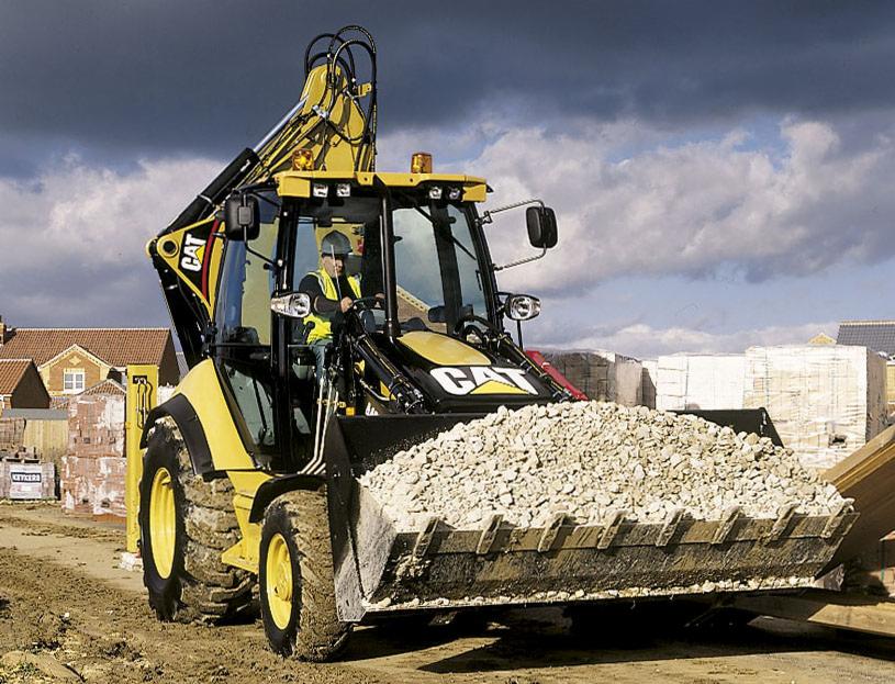 Żółto-czarna koparko-ładowarka CAT z pełną łyżką gruzu na placu budowy, operator w kabinie, w tle budynki w trakcie budowy i pochmurne niebo.