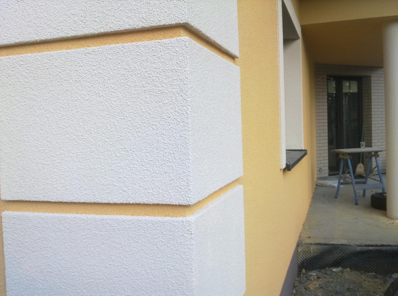 Fragment elewacji domu z dekoracyjnymi, białymi elementami o fakturze baranka, oddzielonymi od reszty żółtej elewacji poziomymi pasami; widoczne okno i fragment tarasu z narzędziami.