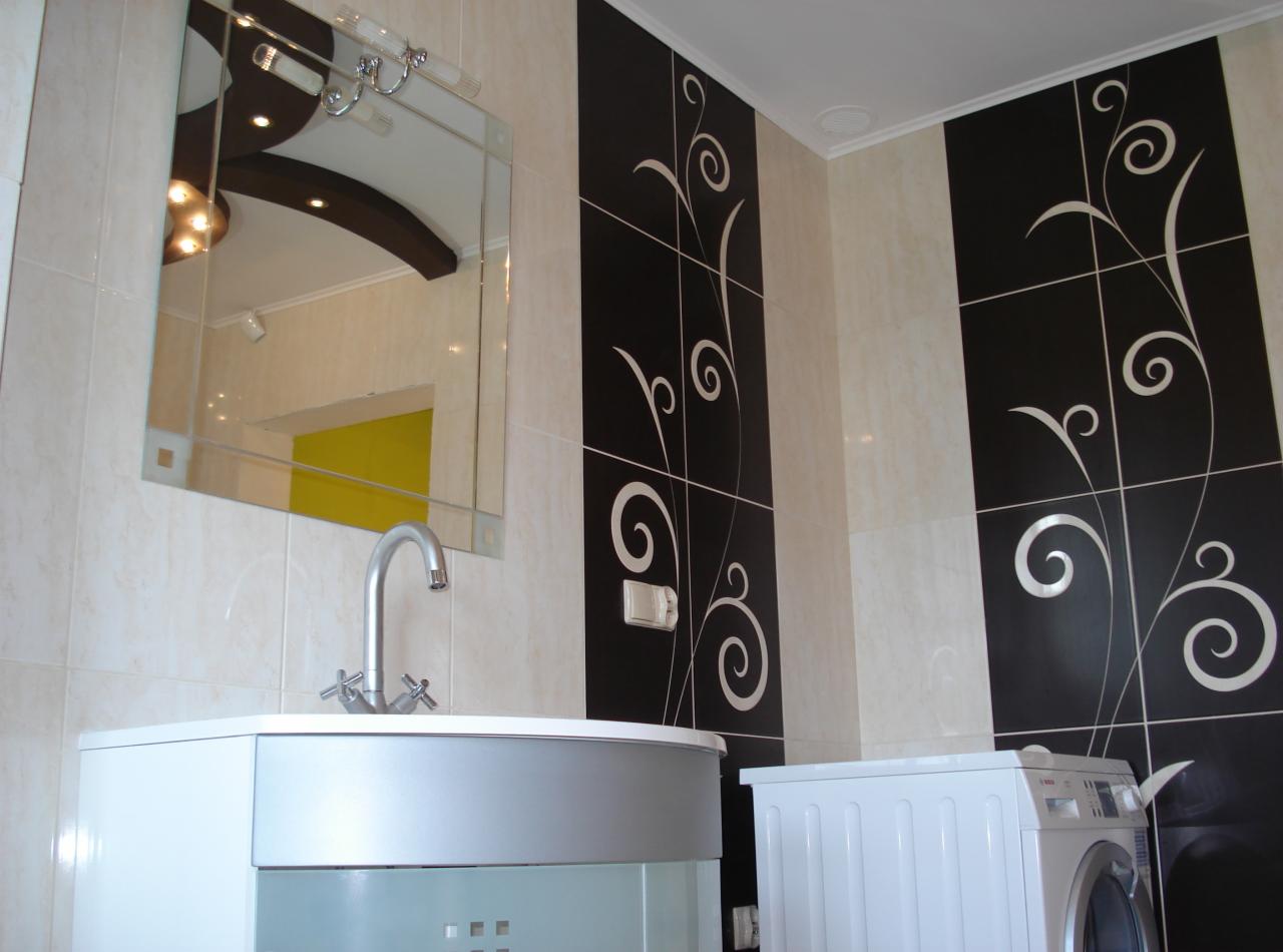 Nowoczesna łazienka z umywalką, lustrem i pralką. Ściany wyłożone białymi i czarnymi płytkami z motywem roślinnym. Widoczne elementy oświetlenia.