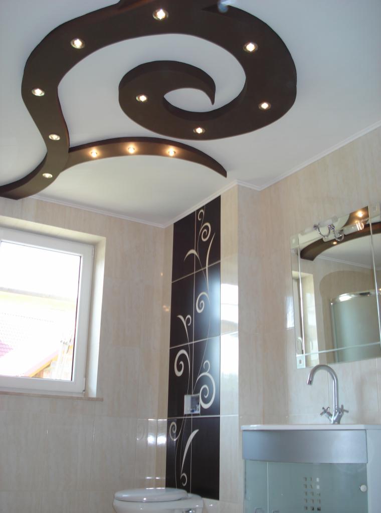 Wykończona łazienka z beżowymi płytkami, dekoracyjnym sufitem podwieszanym z oświetleniem punktowym i czarnym panelem z motywem roślinnym na ścianie.