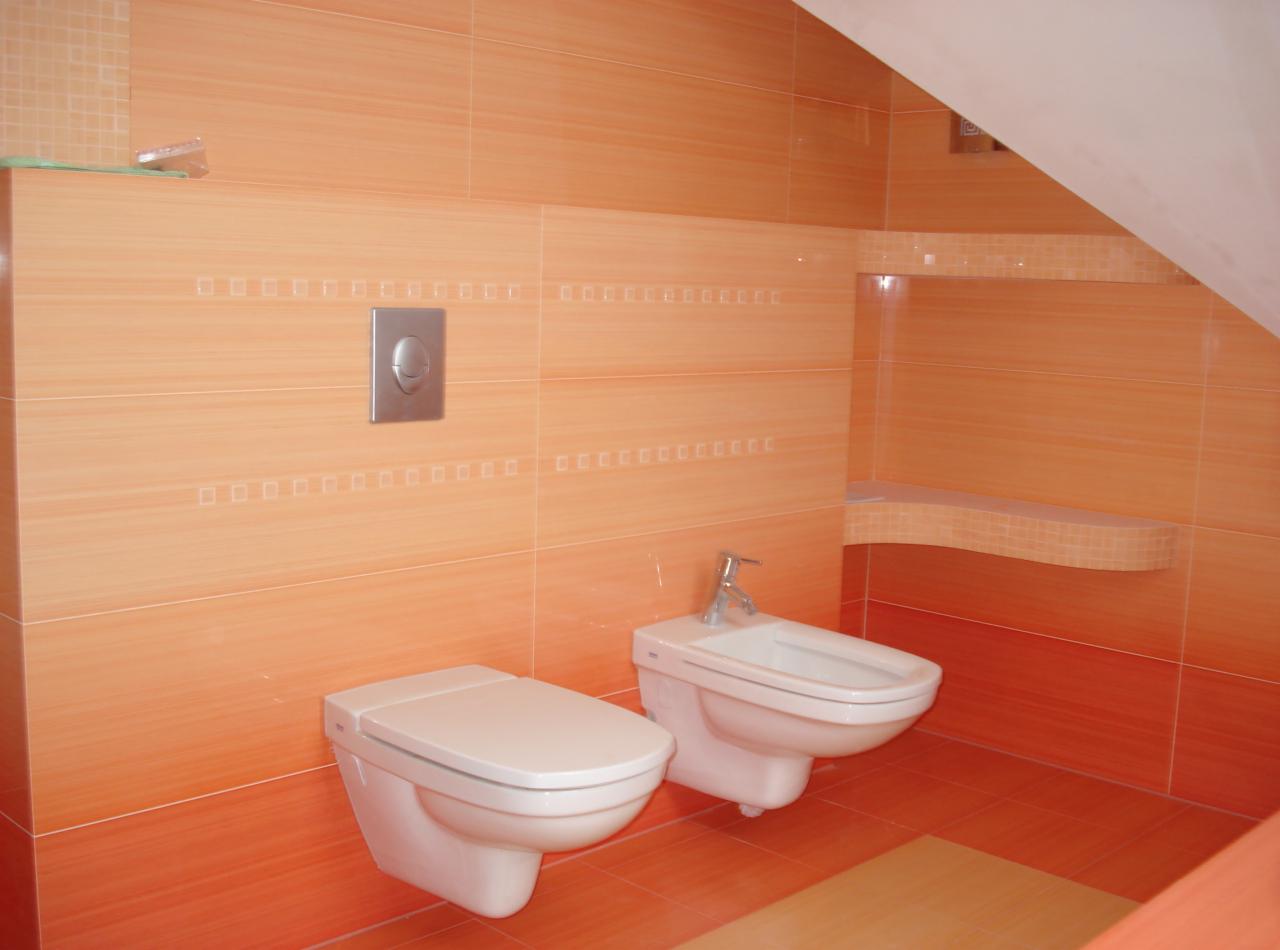 Wykończona łazienka z pomarańczowymi płytkami, wiszącą toaletą i bidetem. Uwagę zwraca półka wnękowa wykończona mozaiką i zabudowa pod skosem.