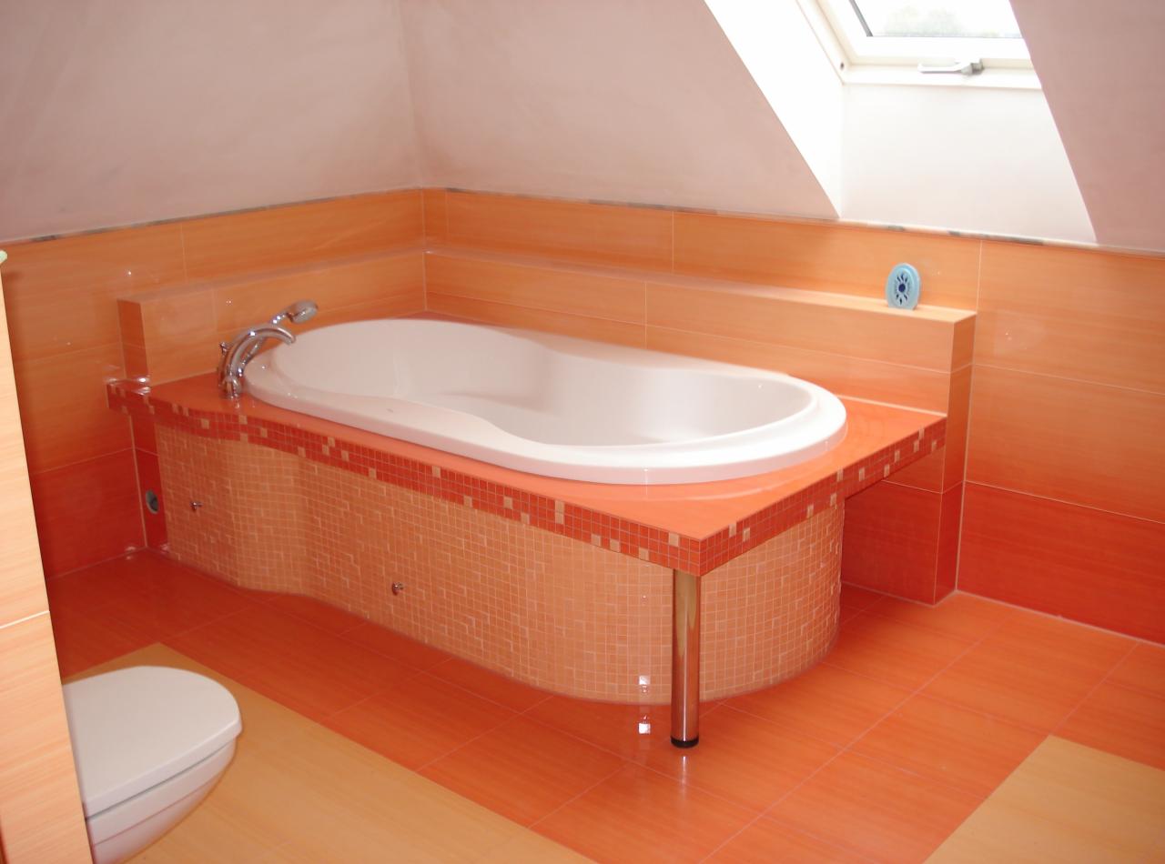 Pomarańczowa łazienka na poddaszu z owalną wanną na podeście wykończonym mozaiką. Widoczne chromowane nóżki i bateria. U góry okno dachowe.