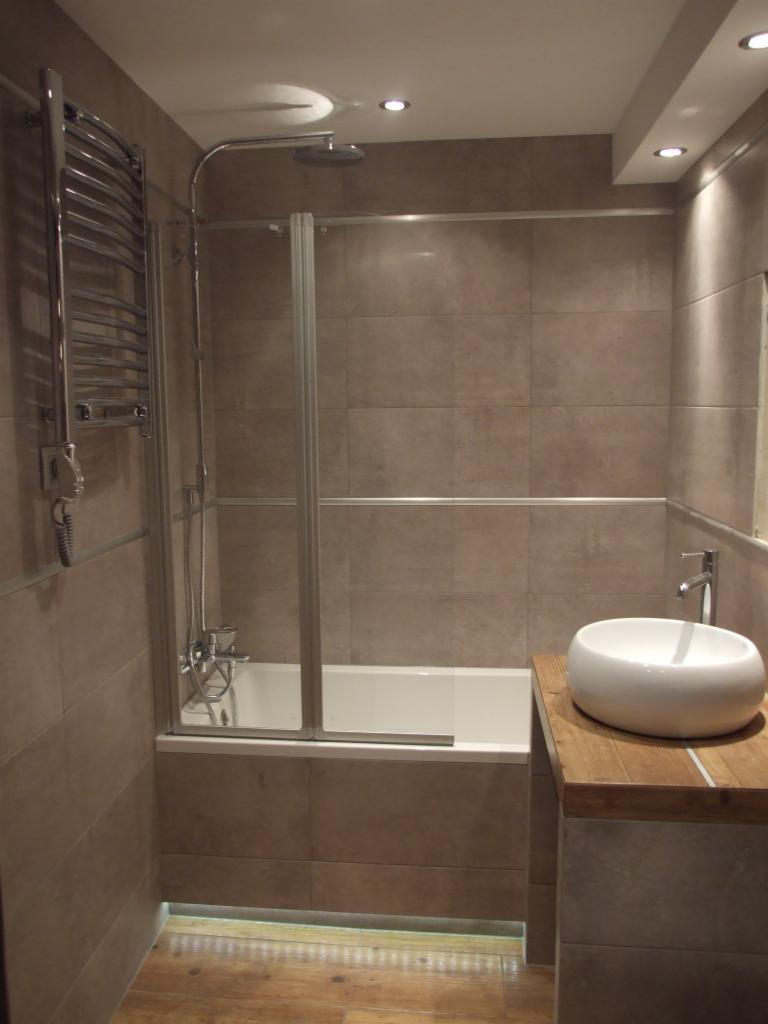 Nowoczesna łazienka z beżowymi płytkami, białą wanną z parawanem i umywalką nablatową na drewnianym blacie. Grzejnik drabinkowy i oświetlenie punktowe.