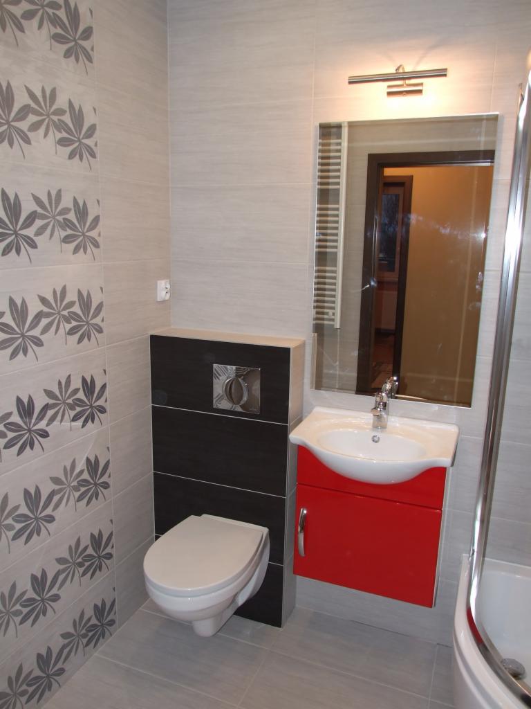 Nowoczesna łazienka z wiszącą toaletą, czerwoną szafką pod umywalką i lustrem. Na ścianach płytki z motywem liści i szare płytki imitujące kamień.