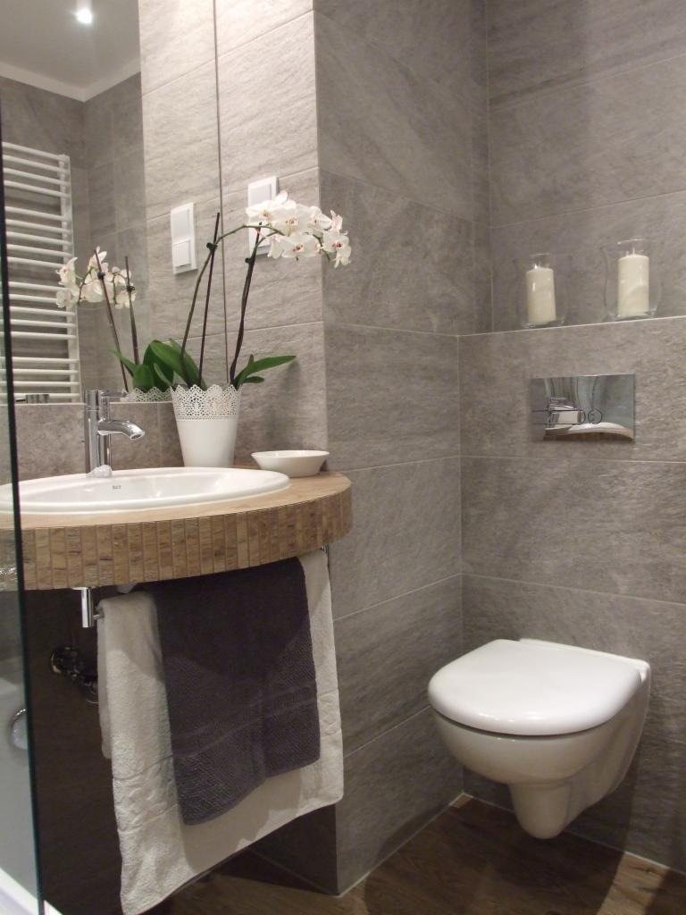 Nowoczesna łazienka z szarymi płytkami, wiszącą toaletą i umywalką z blatem w mozaikę. Biały storczyk w doniczce dodaje elegancji. Widoczne ręczniki i świeczki.