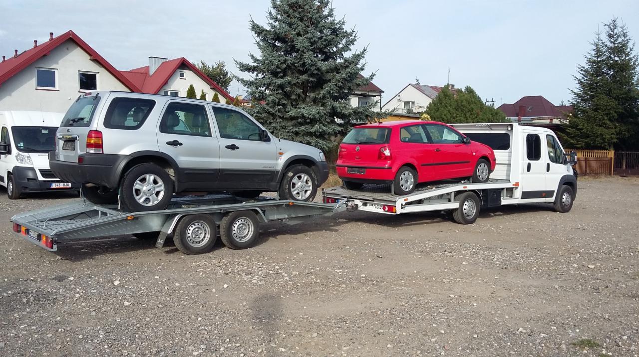 Laweta z dwoma autami: srebrnym SUV-em i czerwonym hatchbackiem, na tle budynków z czerwonymi dachami i zielonego drzewa iglastego. Dzienny kadr.