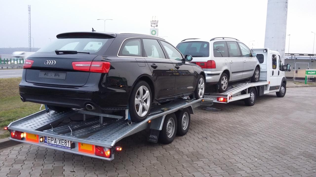 Laweta z dwoma samochodami osobowymi, czarnym Audi A6 i srebrnym vanem, na parkingu z kostki. Widoczny znak drogowy w tle.