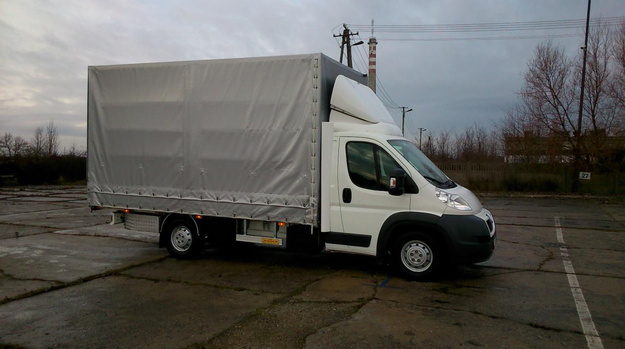 Biały bus z plandeką na parkingu w Ksawerowie, widok z boku. Szara plandeka, biała kabina, szare niebo w tle. Pojazd gotowy do transportu towarów.
