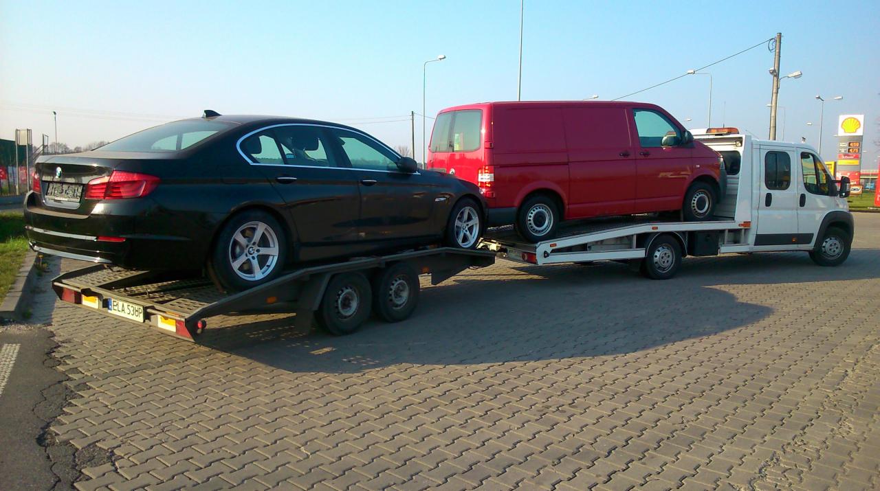 Laweta przewozi czarne auto i czerwony bus na tle stacji benzynowej. Dwa pojazdy na platformie, transport drogowy, widok z boku, kostka brukowa.