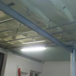 Poddasze po ociepleniu pianką PUR, widoczne metalowe profile i warstwa izolacji natryskowej. Oświetlenie jarzeniowe. Fragment ściany i otwartego przejścia.