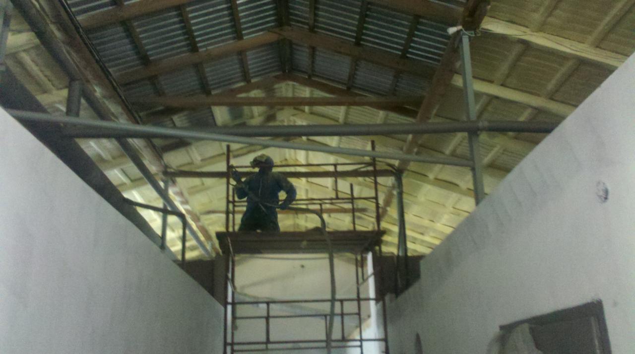 Pracownik w kombinezonie na rusztowaniu, aplikujący piankę izolacyjną na poddasze z blachy falistej. Widoczne ściany boczne i konstrukcja dachu.