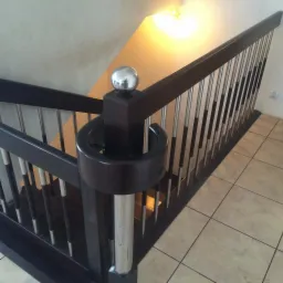 Nowoczesna balustrada schodowa z ciemnego drewna i metalowymi prętami, zwieńczona chromowaną kulą, na tle beżowych płytek podłogowych.