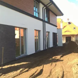 Nowoczesny dom z fragmentami elewacji wykończonymi kamieniem, widoczne ślady po pracach ziemnych i montażu ogrodzenia, w tle pracownik z łopatą.