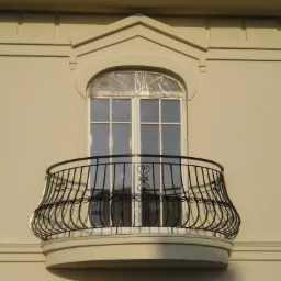 Elegancki balkon z dekoracyjną, czarną, metalową balustradą na tle kremowej fasady budynku z oknem zabezpieczonym folią.