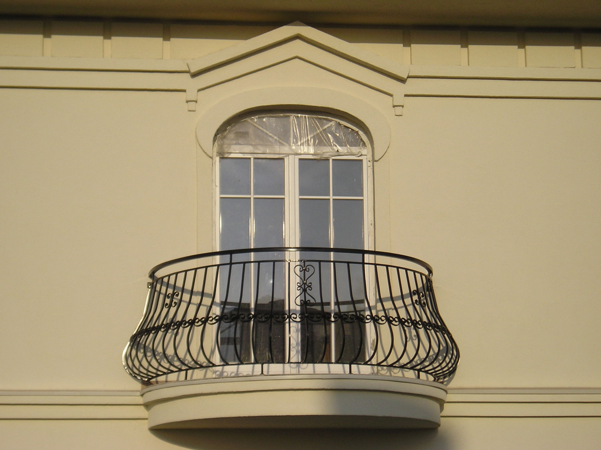 Elegancki balkon z dekoracyjną, czarną, metalową balustradą na tle kremowej fasady budynku z oknem zabezpieczonym folią.