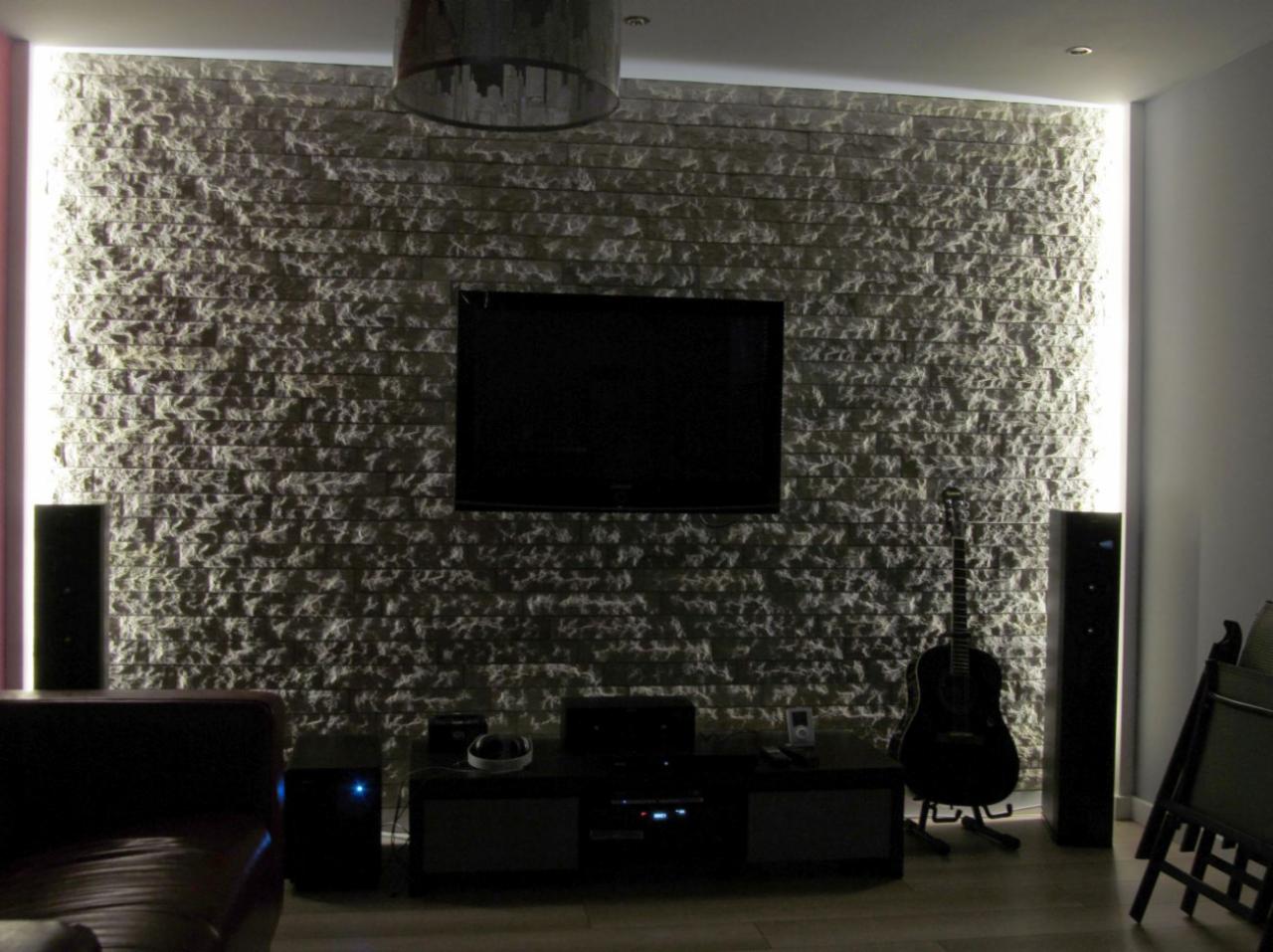 Ściana w salonie wyłożona kamiennymi płytkami imitującymi naturalny kamień, podświetlona po bokach listwami LED, z wiszącym telewizorem i gitarą opartą o ścianę.