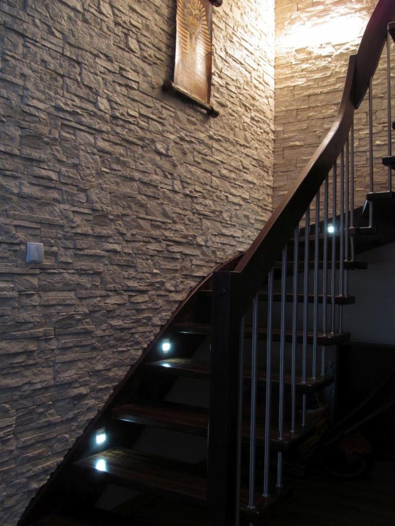 Ściana wykończona okładziną elewacyjną imitującą kamień, z drewnianymi schodami z podświetleniem LED i obrazkiem w stylu egipskim.