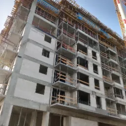 Wielopiętrowy budynek w stanie surowym, widoczne szalunki i zbrojenia balkonów, jasne bloczki konstrukcyjne, w tle żuraw budowlany.