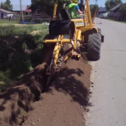 Żółty traktor Vermeer kopie wąski rów wzdłuż asfaltowej drogi, operator w kamizelce odblaskowej, w tle zabudowania wiejskie i częściowo zachmurzone niebo.
