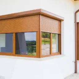 Kanciaste okno z brązowymi roletami zewnętrznymi w kolorze drewna, zamontowanymi na białej ścianie domu. Widoczne dwa okna, jedno kanciaste, drugie zaokrąglone, z refleksami otoczenia w szybach.