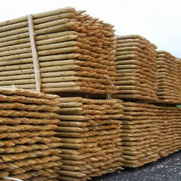 Stosy impregnowanych, zaostrzonych pali drewnianych, spięte białymi taśmami, składowane na żwirowym podłożu, z fragmentem torów kolejowych w tle.