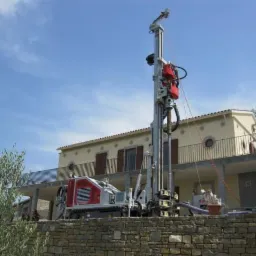 Wiertnica geologiczna na tle domu z balkonem i błękitnego nieba. Urządzenie w kolorach szarości, bieli i czerwieni. Prace wiertnicze na zewnątrz budynku.