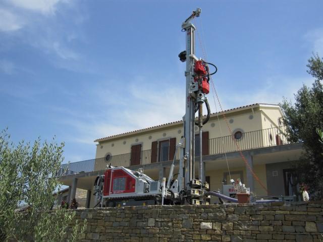 Wiertnica geologiczna na tle domu z balkonem i błękitnego nieba. Urządzenie w kolorach szarości, bieli i czerwieni. Prace wiertnicze na zewnątrz budynku.