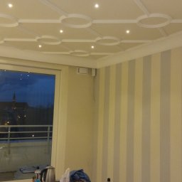 RAFAŁ POŚLEDNIAK BUD-RAF - Pomalowane ściany w pionowe pasy, ozdobny sufit z oświetleniem punktowym. Widok z okna na miasto nocą. Fragment drabiny malarskiej w prawym dolnym rogu.