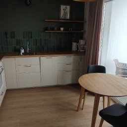 RAFAŁ POŚLEDNIAK BUD-RAF - Jasna kuchnia z ciemnozieloną ścianą i płytkami, drewnianym blatem i stołem. Widok na balkon z krzesłami i budynki w tle. Nowoczesne wykończenie wnętrza.