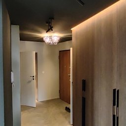 RAFAŁ POŚLEDNIAK BUD-RAF - Nowoczesny przedpokój z ciemnym sufitem, oświetleniem LED i szafą w zabudowie z długimi, czarnymi uchwytami. W tle widoczne drzwi i odkurzacz.