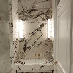 RAFAŁ POŚLEDNIAK BUD-RAF - Elegancka łazienka z marmurowymi ścianami i blatem, oświetlona pionowymi lampami. Złote detale armatury i białe drzwi dodają luksusowego charakteru.