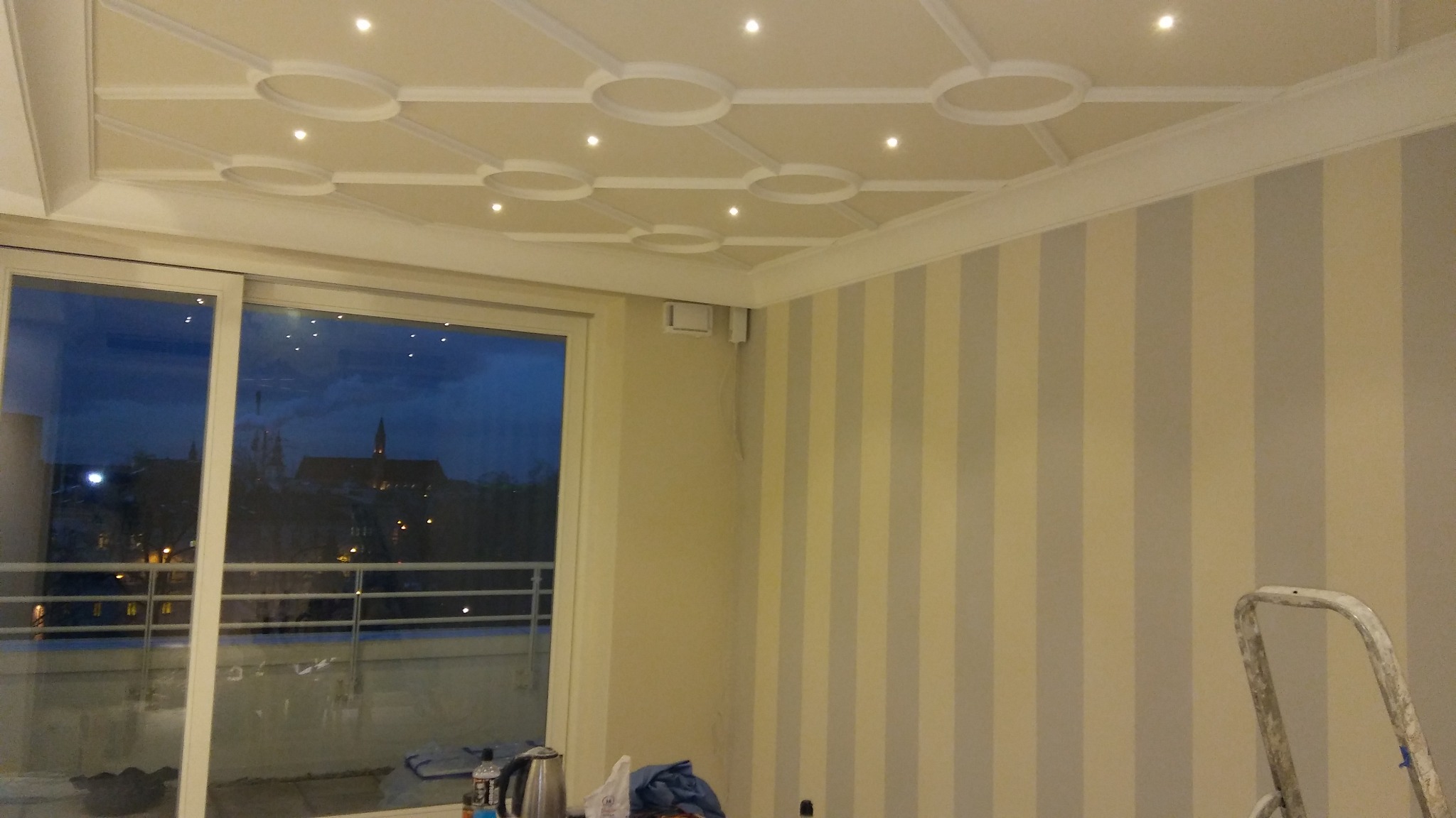 Pomalowane ściany w pionowe pasy, ozdobny sufit z oświetleniem punktowym. Widok z okna na miasto nocą. Fragment drabiny malarskiej w prawym dolnym rogu.