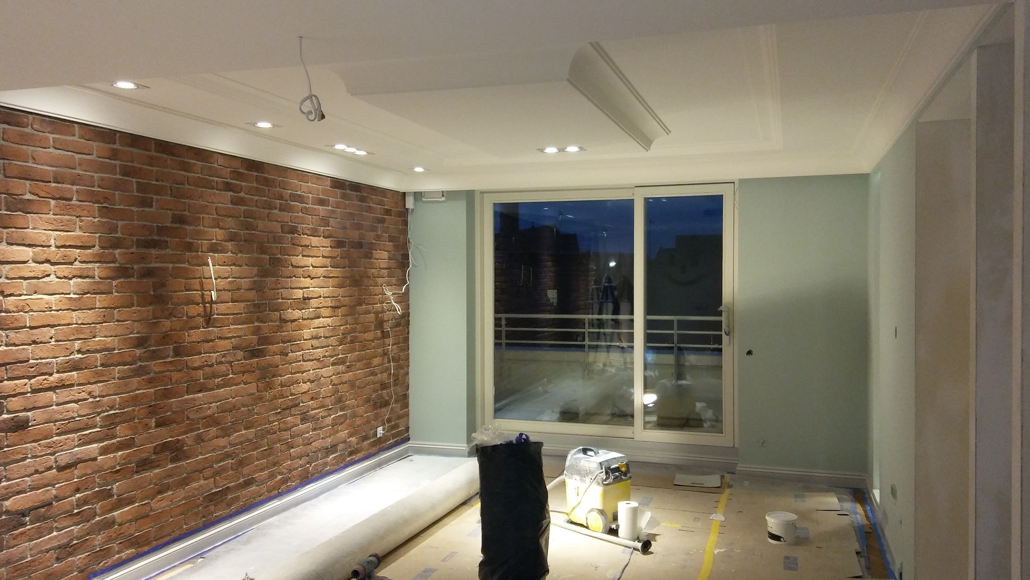 Pokój w trakcie wykańczania: ściana z cegły, pomalowane na jasno ściany, okno balkonowe. Widoczne narzędzia i materiały budowlane. Sufit z dekoracyjnymi elementami.