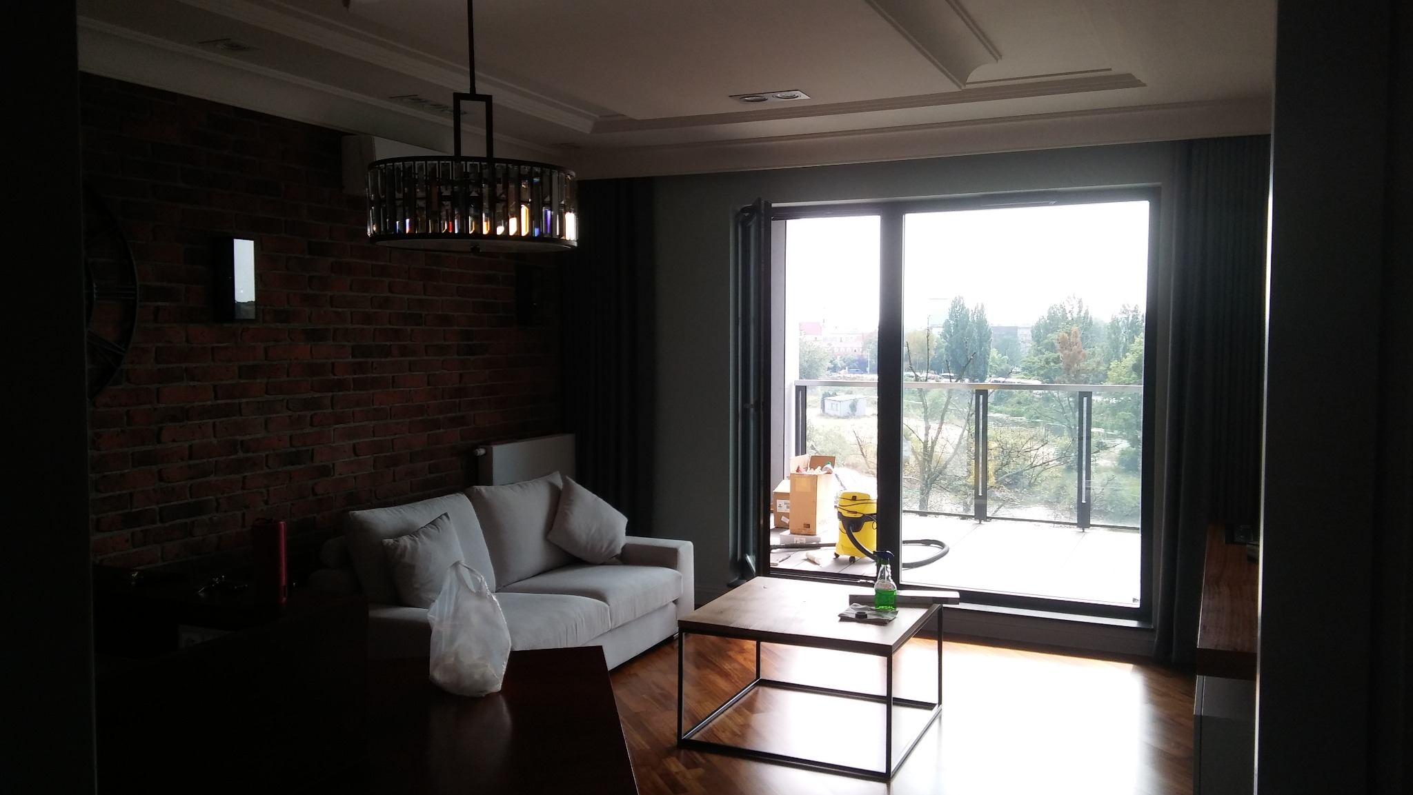 Nowoczesny salon z ceglaną ścianą, szarą sofą i widokiem na balkon. Na balkonie widoczne kartony i odkurzacz, sugerujące prace wykończeniowe.