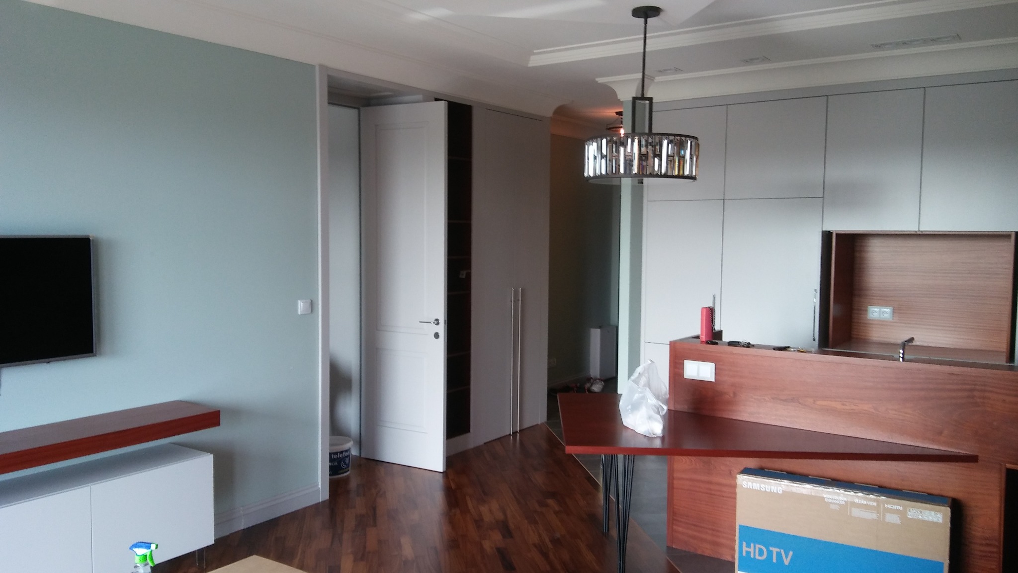 Nowoczesne wnętrze po remoncie: błękitna ściana, zabudowa kuchenna z drewnianymi akcentami, białe drzwi do garderoby i parkiet. Widoczny karton po telewizorze.