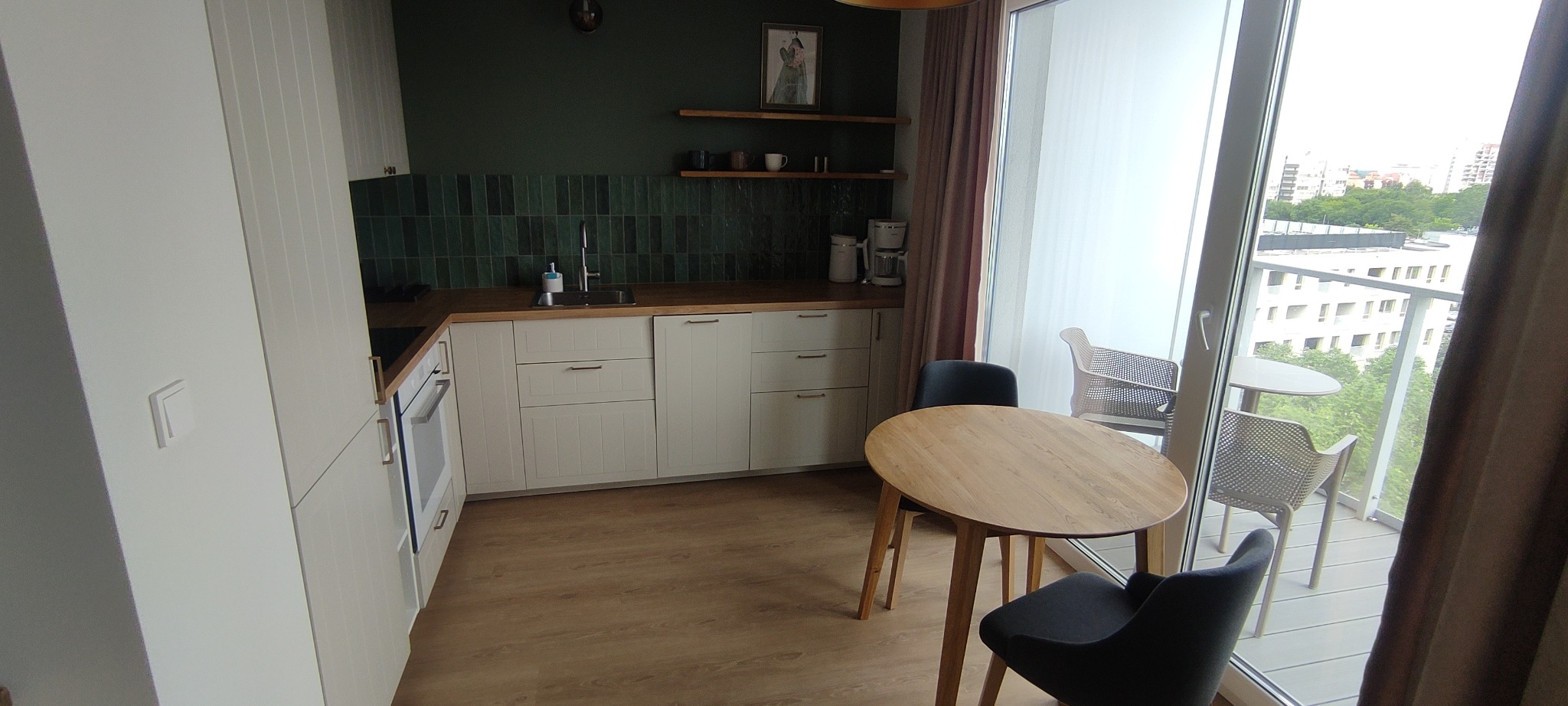 Jasna kuchnia z ciemnozieloną ścianą i płytkami, drewnianym blatem i stołem. Widok na balkon z krzesłami i budynki w tle. Nowoczesne wykończenie wnętrza.