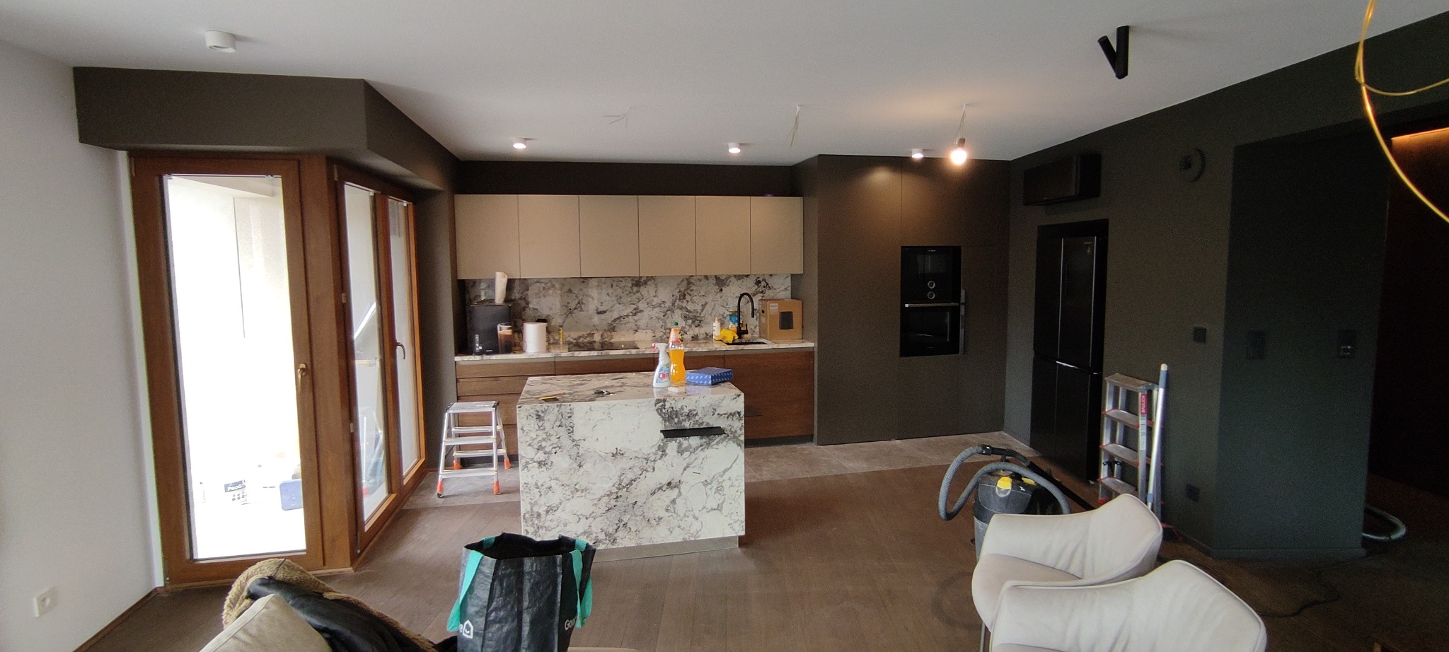 Nowoczesna kuchnia w trakcie wykańczania: marmurowa wyspa, beżowe szafki, ciemne ściany. Widoczne narzędzia i akcesoria, sugerujące etap po remoncie, przed ostatecznym sprzątaniem.