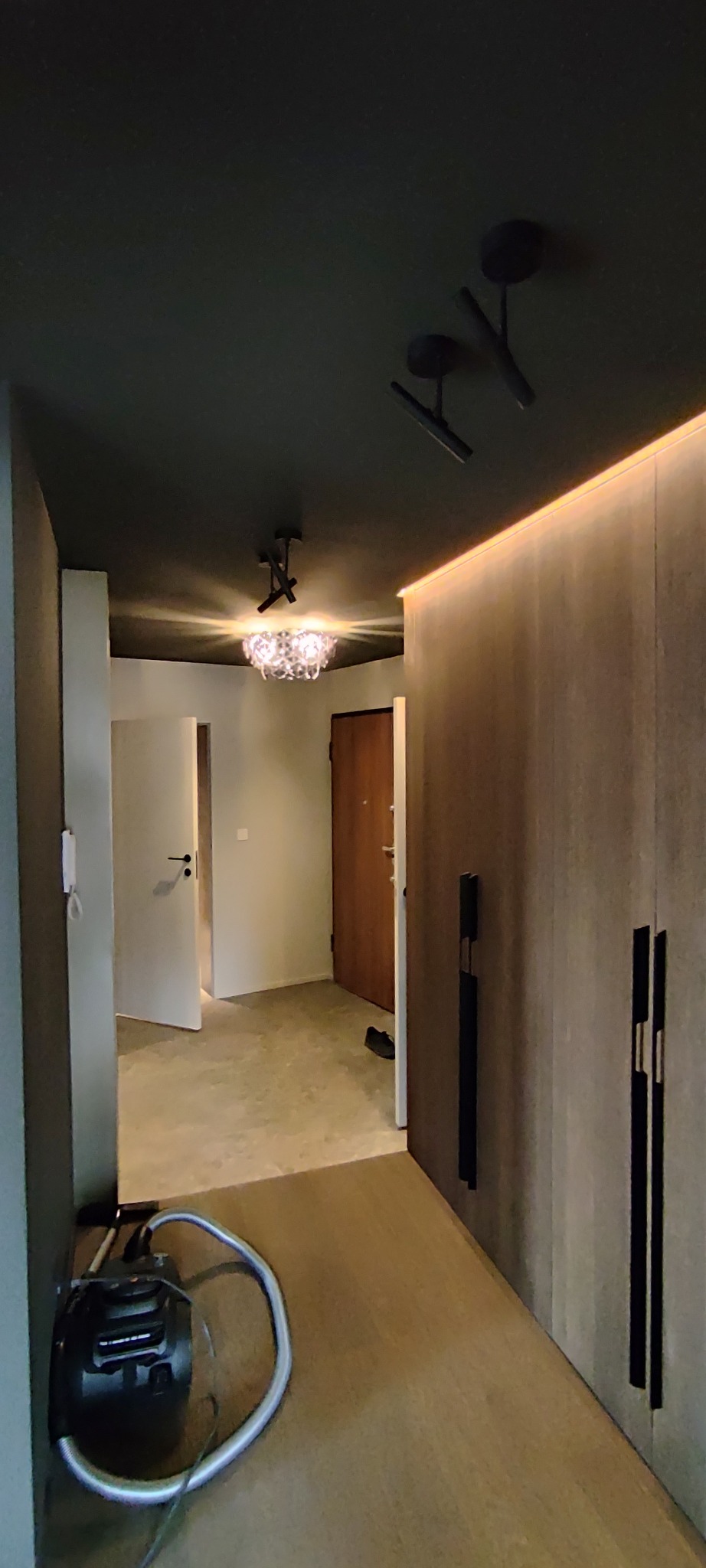 Nowoczesny przedpokój z ciemnym sufitem, oświetleniem LED i szafą w zabudowie z długimi, czarnymi uchwytami. W tle widoczne drzwi i odkurzacz.