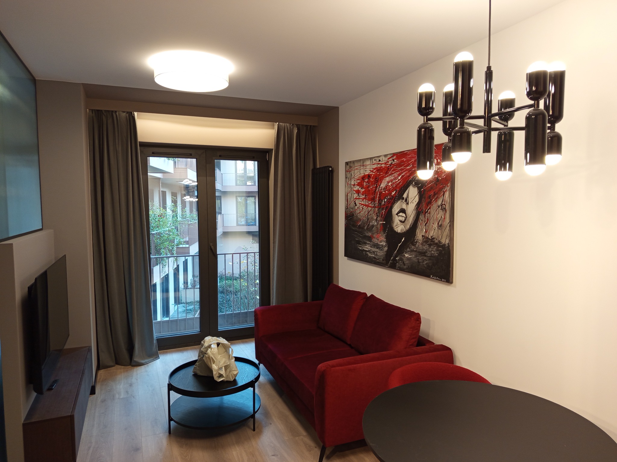 Nowoczesny salon z czerwonymi akcentami: sofa, obraz, i designerska lampa. Widok na balkon przez okno z szarymi zasłonami. Minimalistyczny stolik kawowy i telewizor.