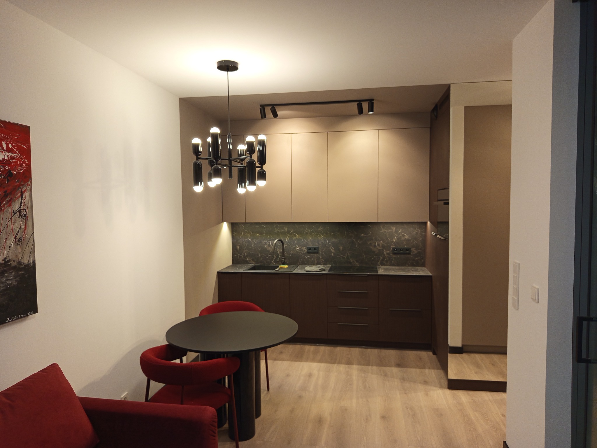 Nowoczesna kuchnia z ciemnymi szafkami, marmurowym blatem i designerską lampą. Czerwone krzesła i okrągły stół dodają akcentu kolorystycznego.
