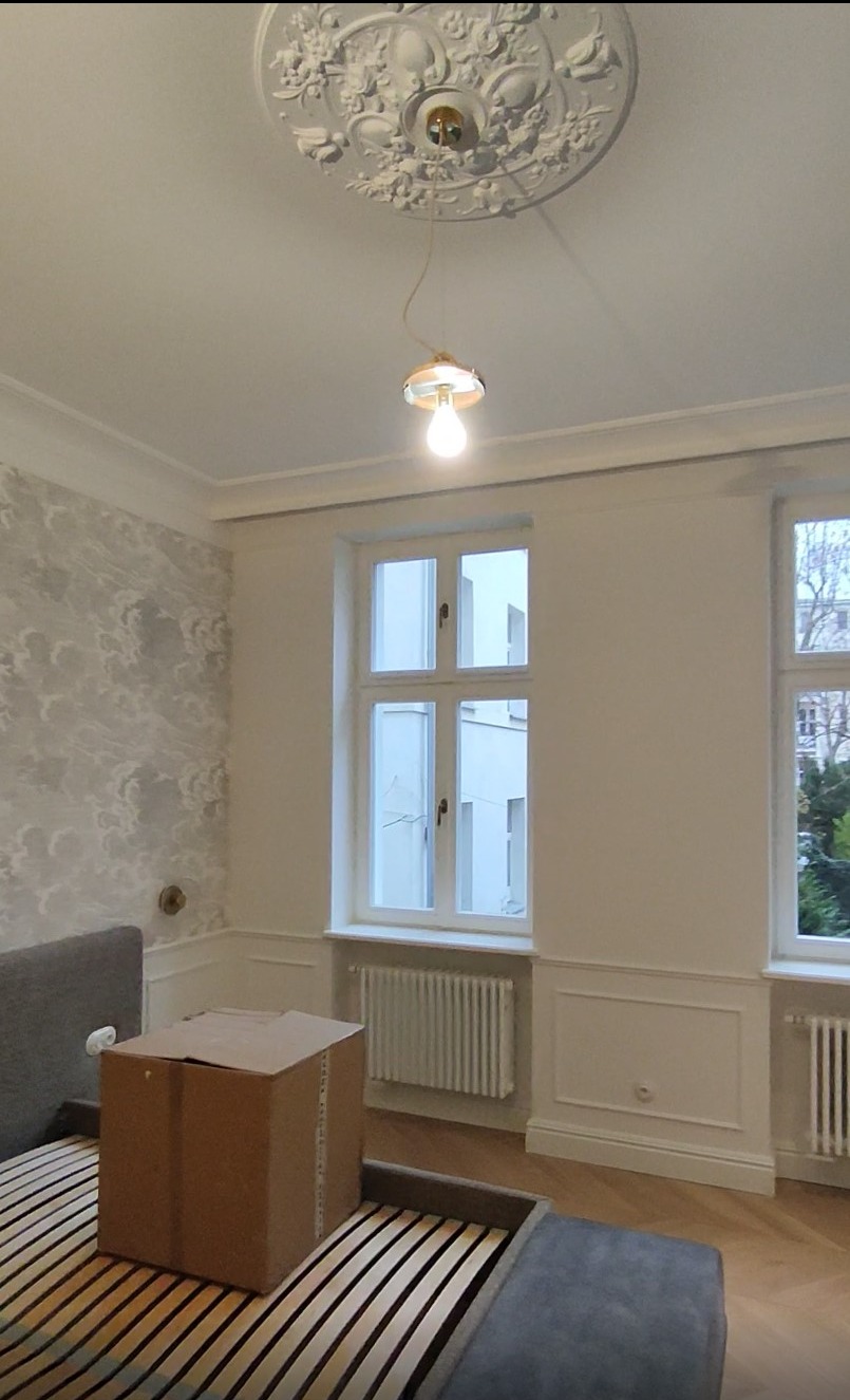 Pokój po remoncie: białe ściany z dekoracyjną sztukaterią, wzorzysta tapeta, drewniana podłoga, okna z widokiem na zieleń, karton na łóżku i ozdobny żyrandol.