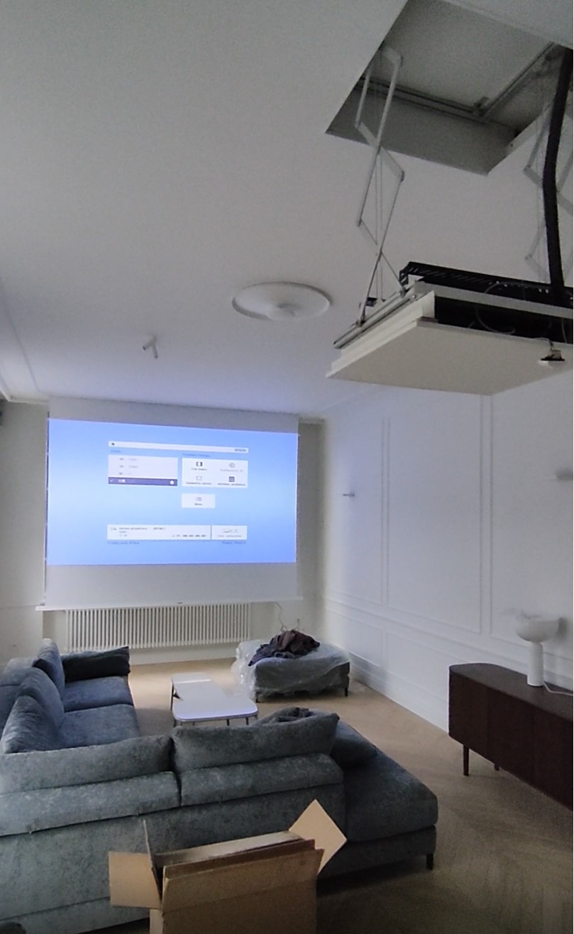 Salon z szarą sofą i ekranem projekcyjnym opuszczonym z sufitu. Widoczne białe ściany z dekoracyjnymi panelami i karton na podłodze.