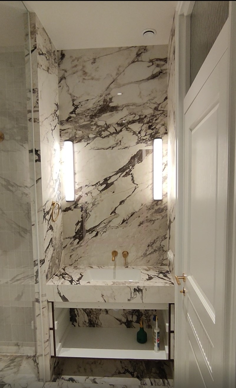 Elegancka łazienka z marmurowymi ścianami i blatem, oświetlona pionowymi lampami. Złote detale armatury i białe drzwi dodają luksusowego charakteru.