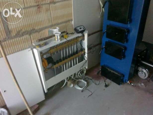 Instalacja ogrzewania podłogowego z rozdzielaczem w szafce i niebieski piec CO. Widoczne przewody i elementy instalacji. Ściana z cegły dziurawki w tle.